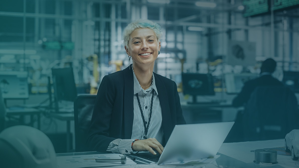 A professional woman working at a desk with a laptop in a modern office environment, featuring blurred background elements that suggest a collaborative workspace.